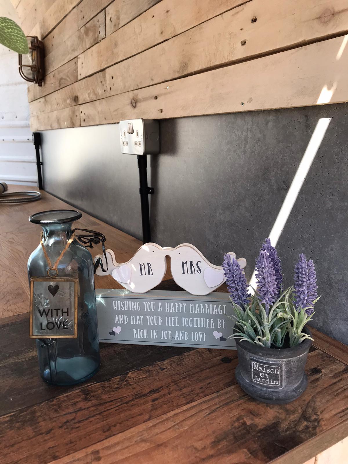 A wooden bar with a bottle that says 'with love' on it, an ornament of two birds marked 'Mr' and 'Mrs' and a small vase of lavender