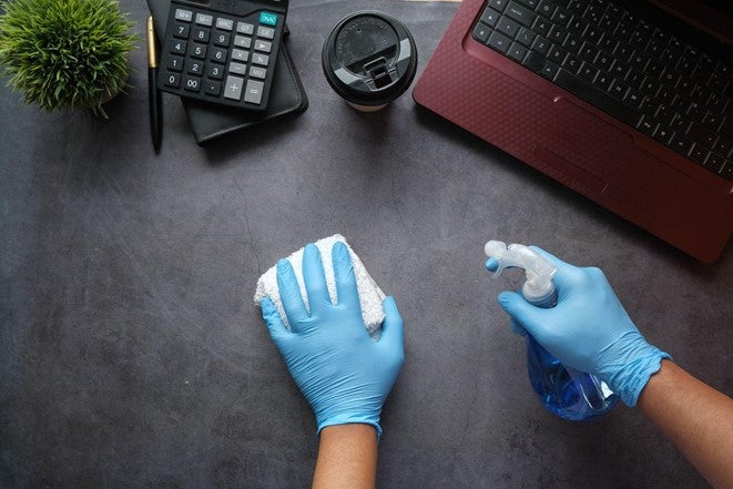A cleaner at an office in Manchester wears blue gloves while using spray detergent and a cloth to clean a desk with a laptop and phone on it