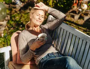 Frau mit Tasse in der Hand liegt in der Sonne auf einer Bank.