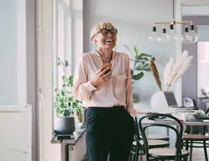 Ältere Frau mit Brille lacht mit Handy in der Hand.