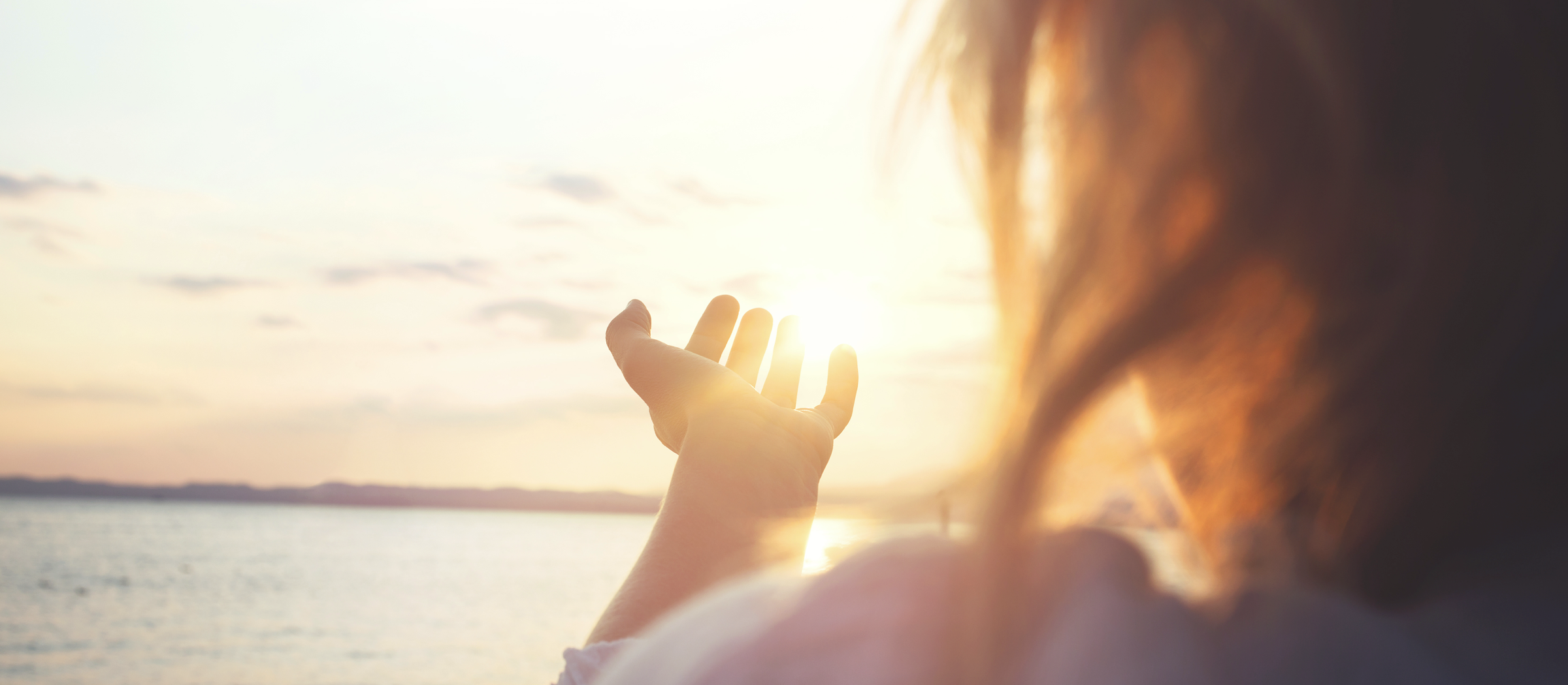 Frau schaut an einem See Richtung Sonne.