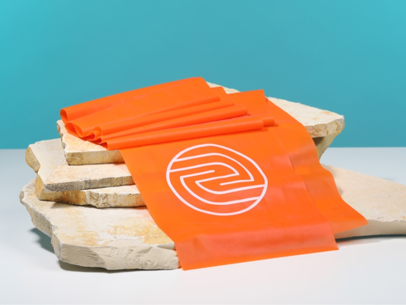 Oranges Theraband mit ENTDECKE DEIN NEO-Logo liegt auf Steinplatten.