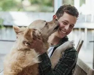 Großer Hund schleckt die Wange seines Herrchens, dieser freut sich und umarmt den Hund.