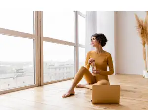 Frau in gelber Yoga-Kleidung sitzt auf dem Boden und schaut aus dem Fenster.