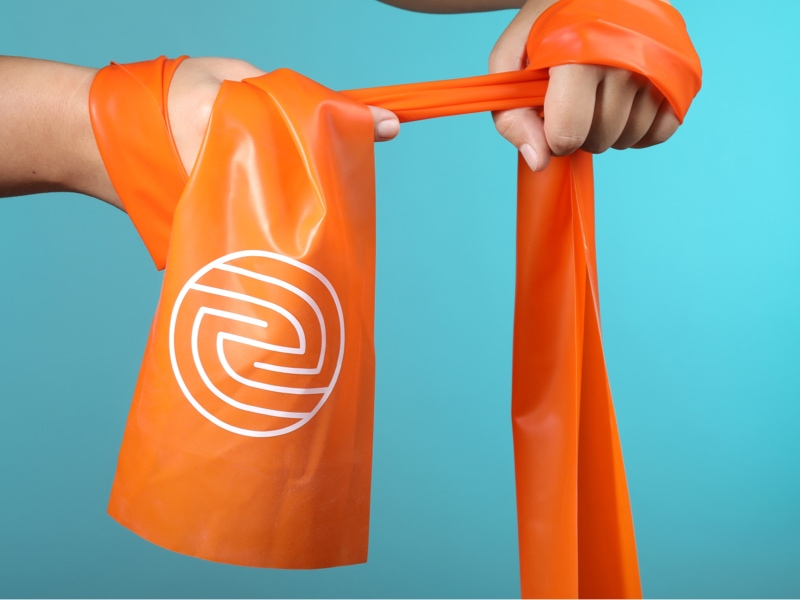 Oranges Theraband mit ENTDECKE DEIN NEO-Logo wird mit zwei Händen gehalten.