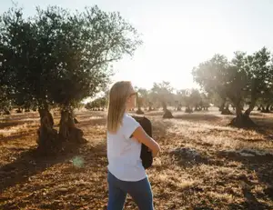 Frau in T-Shirt hält ihren Rucksack und wandert zwischen Olivenbäumen.