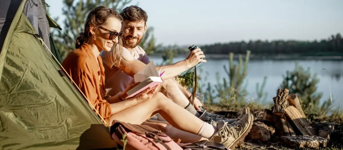 Ein Pärchen zeltet am See und liest in einem Buch.