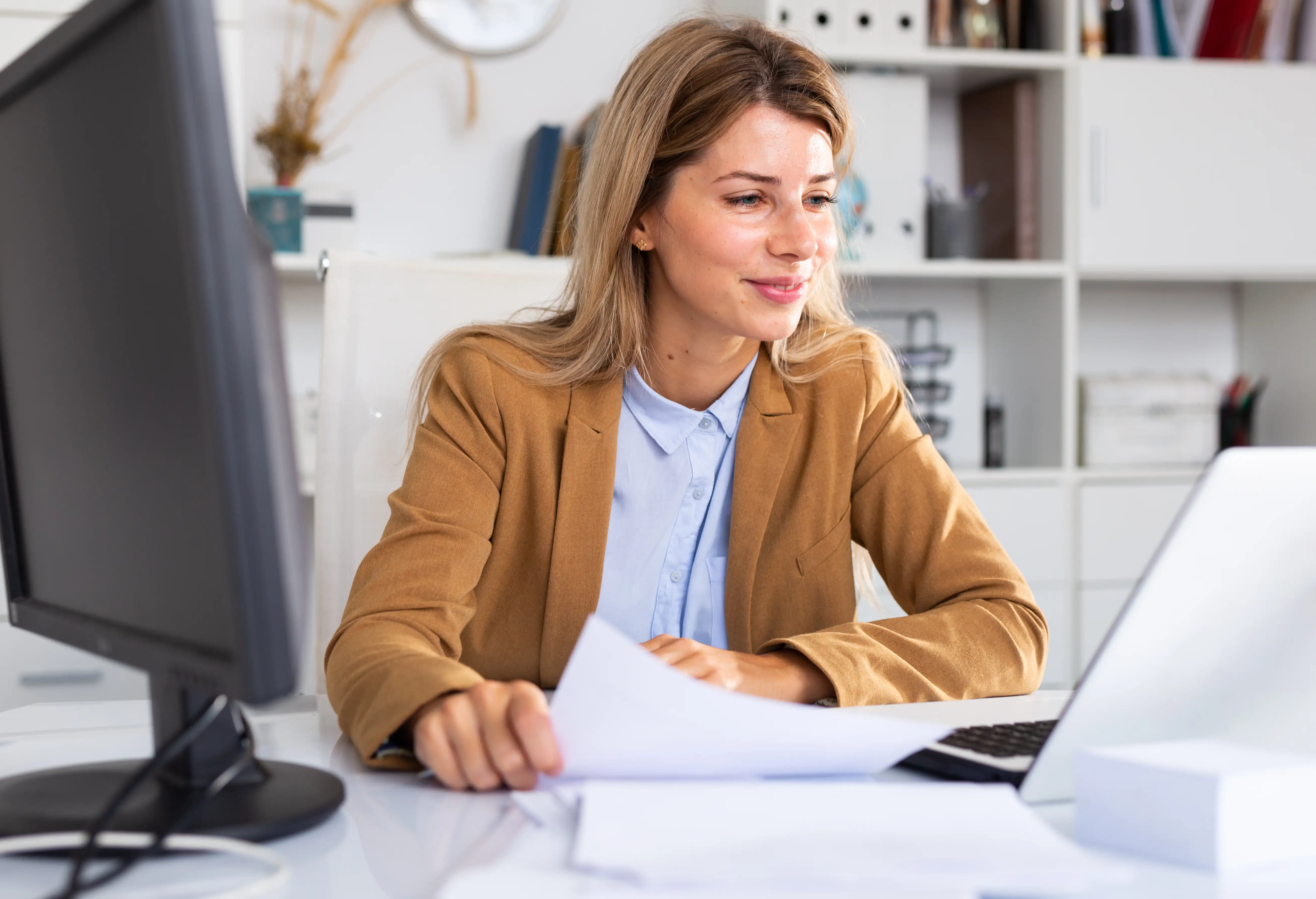 Frau in orangenem Blazer sitzt am Schreibtisch und schaut auf ihren Laptop.