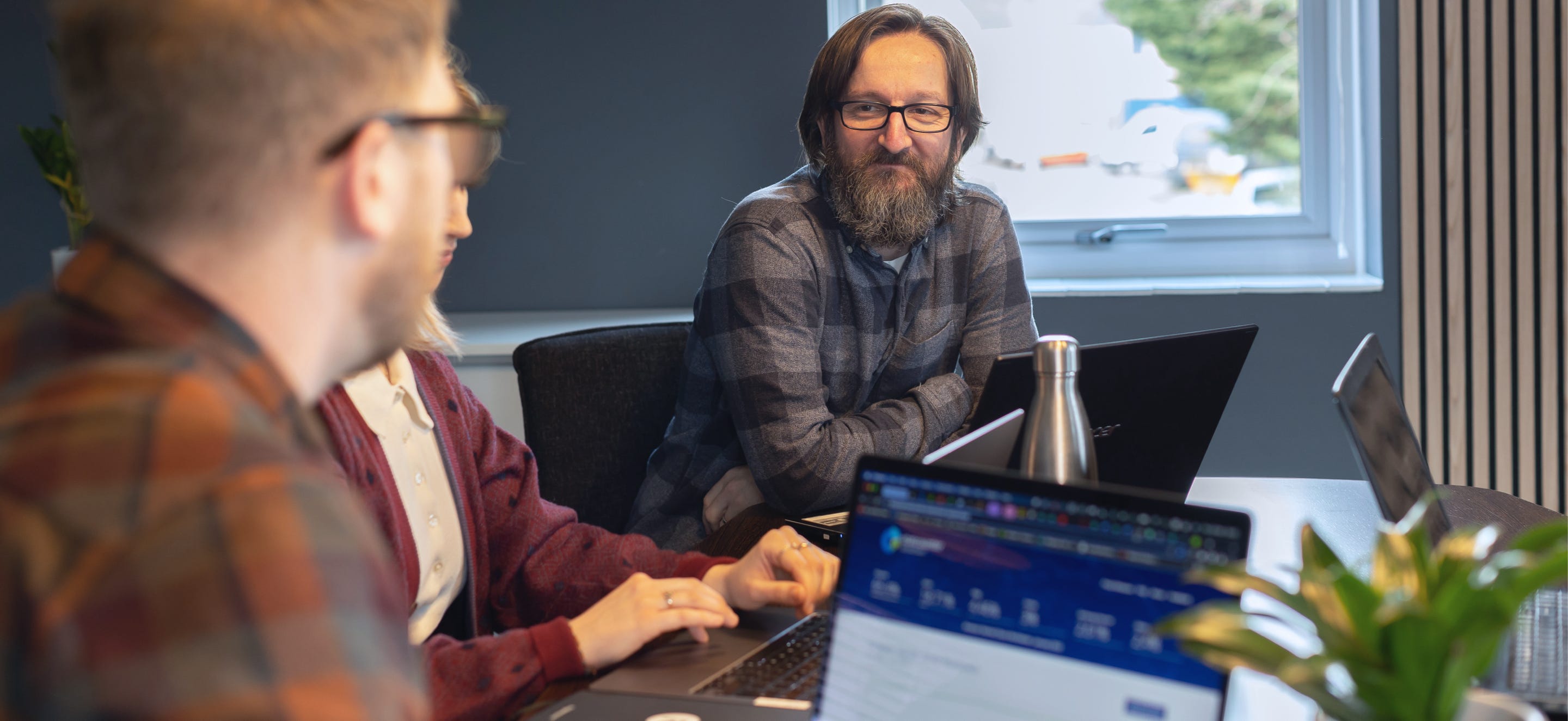 Two men and lady in a meeting room, plus laptops and a window in the background