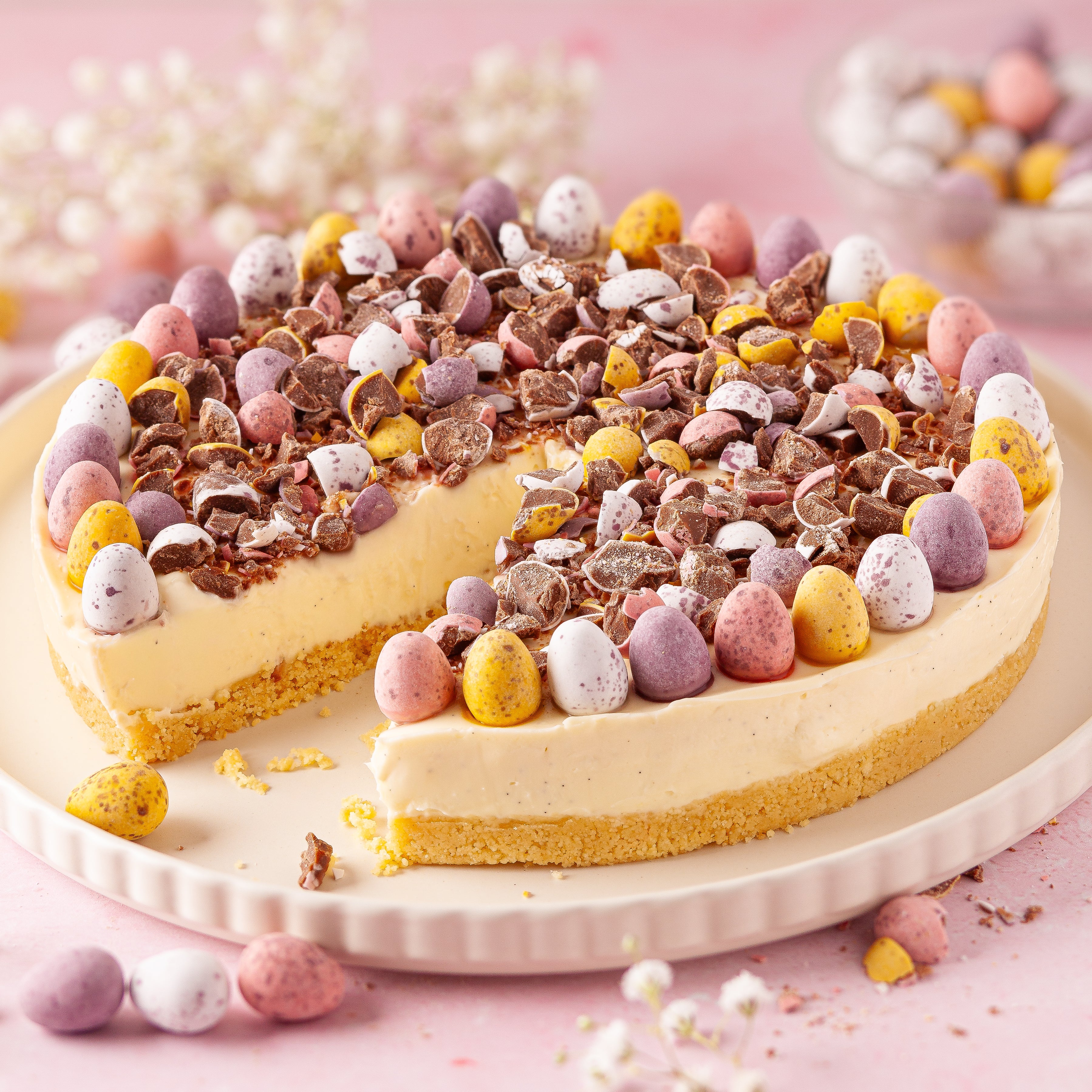 A no bake vanilla mini egg cheesecake with a slice cut out, decorated with whole and crushed mini chocolate eggs.