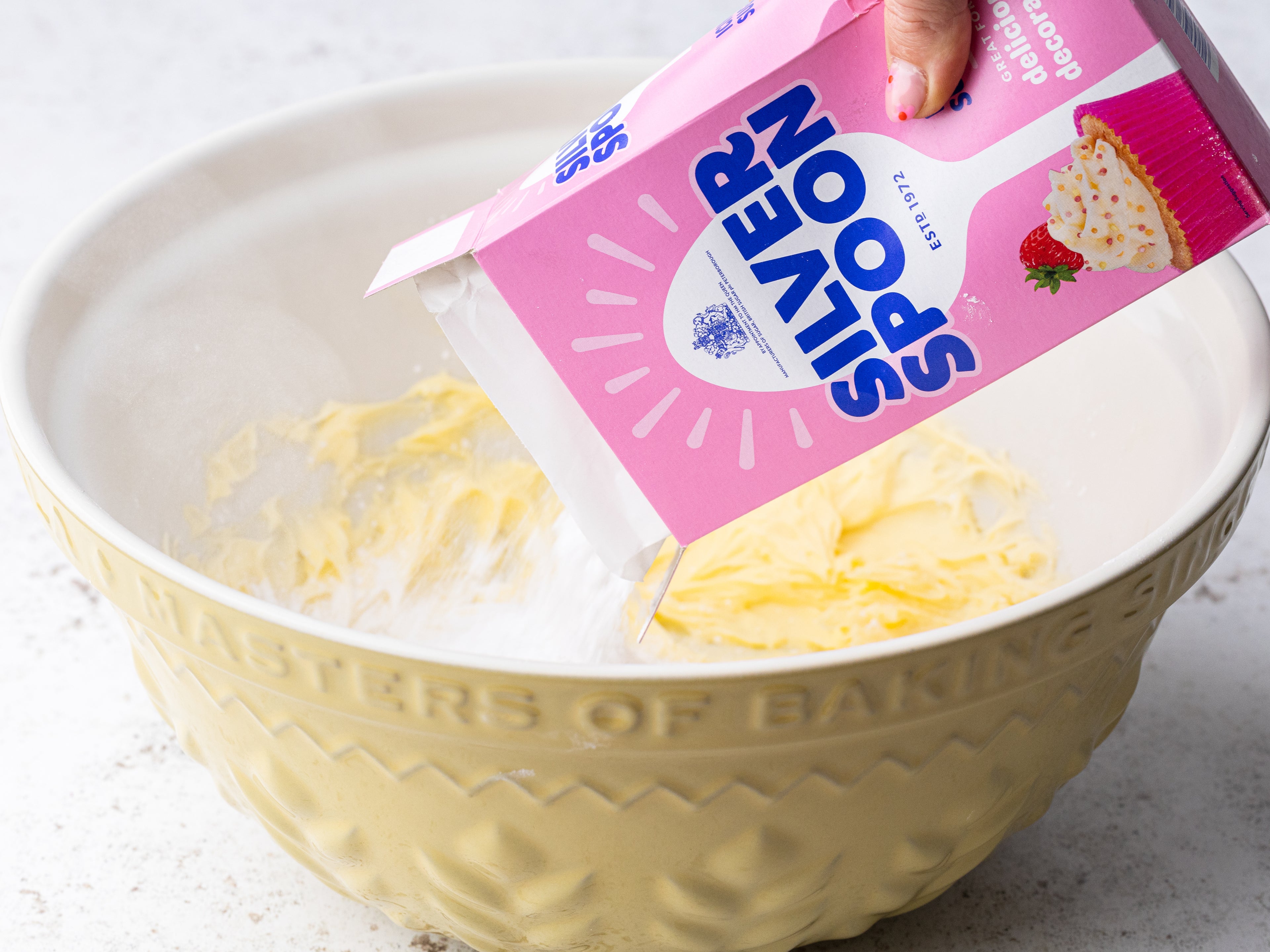 A pink pack of Silver Spoon icing sugar being poured into a large baking bowl that has some cake batter in it.