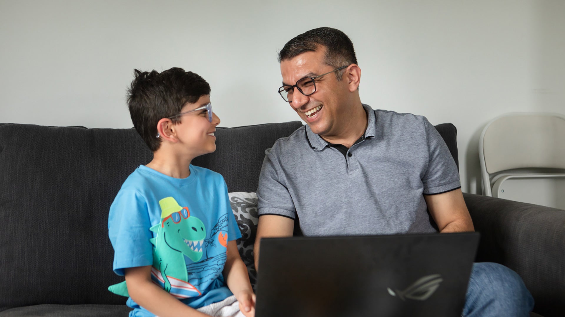 Father and son sitting side by side on a sofa with a laptop in front of them. They are looking at each other and smiling