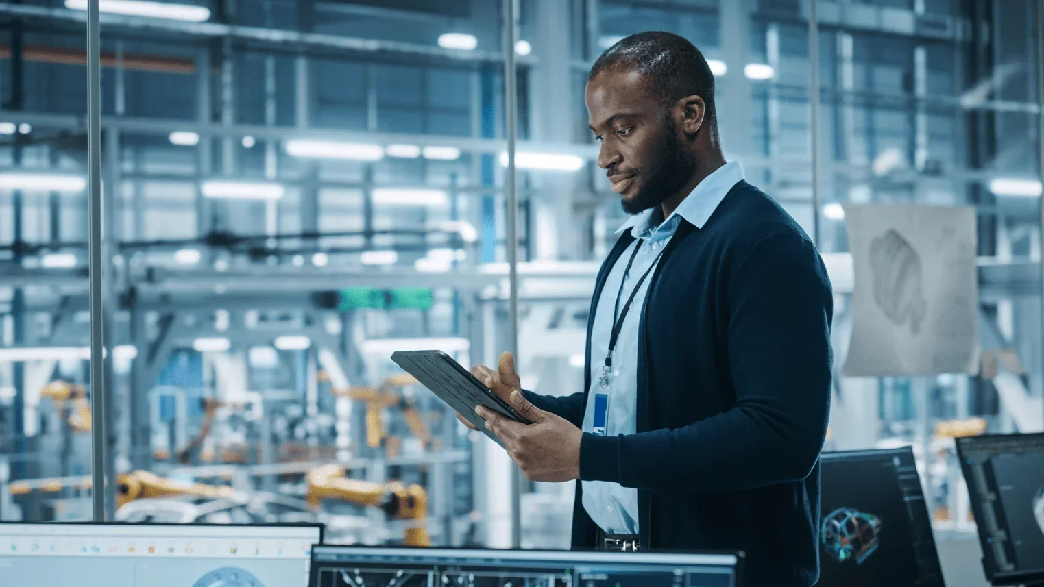 Professional using a tablet to start energy procurement with a broker. He's in a modern industrial facility, with computer monitors in the foreground and automated manufacturing equipment visible through glass in the background.