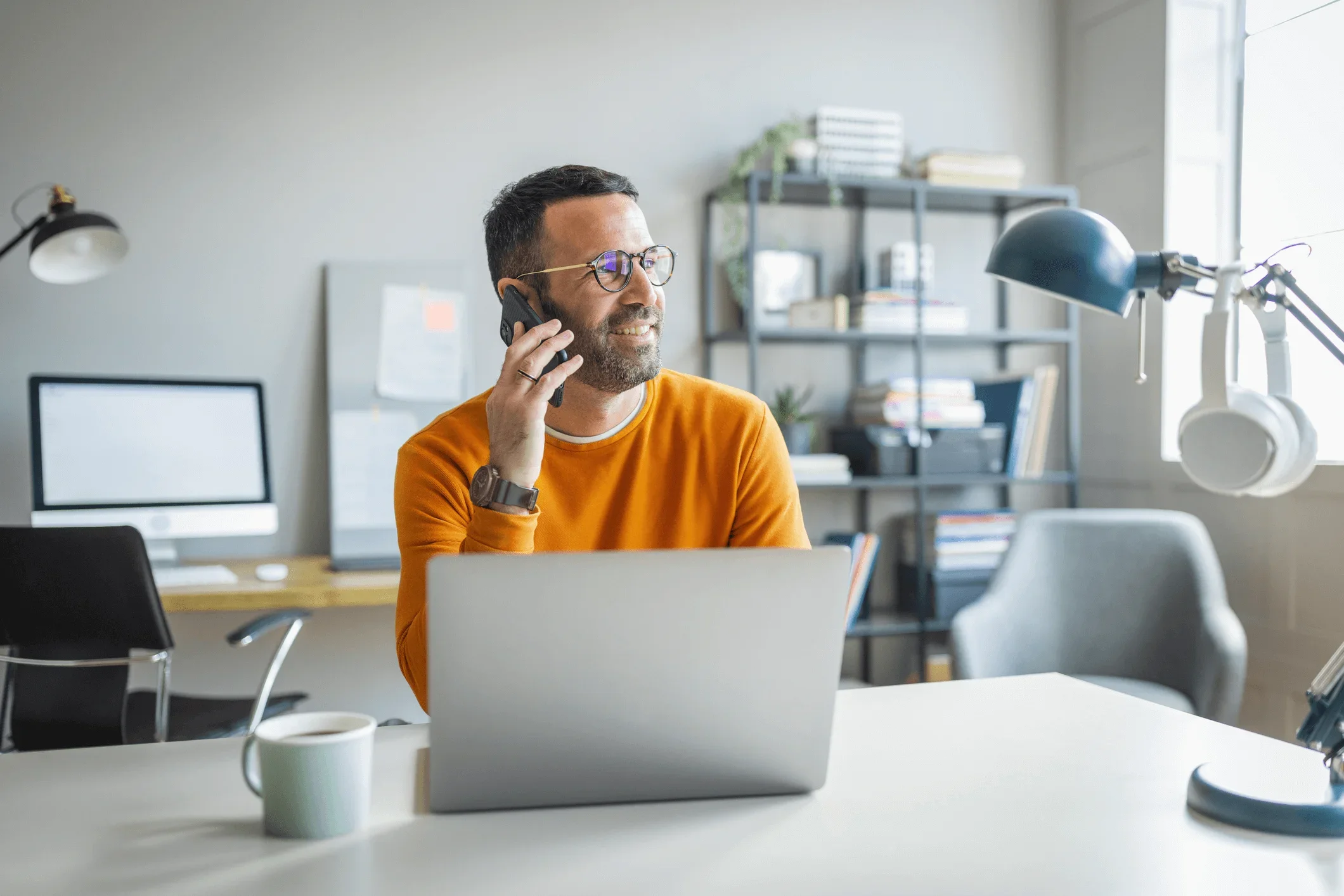 A small business owner sits in his studio with a laptop. He's on his mobile phone speaking to a business energy broker about his next energy contract