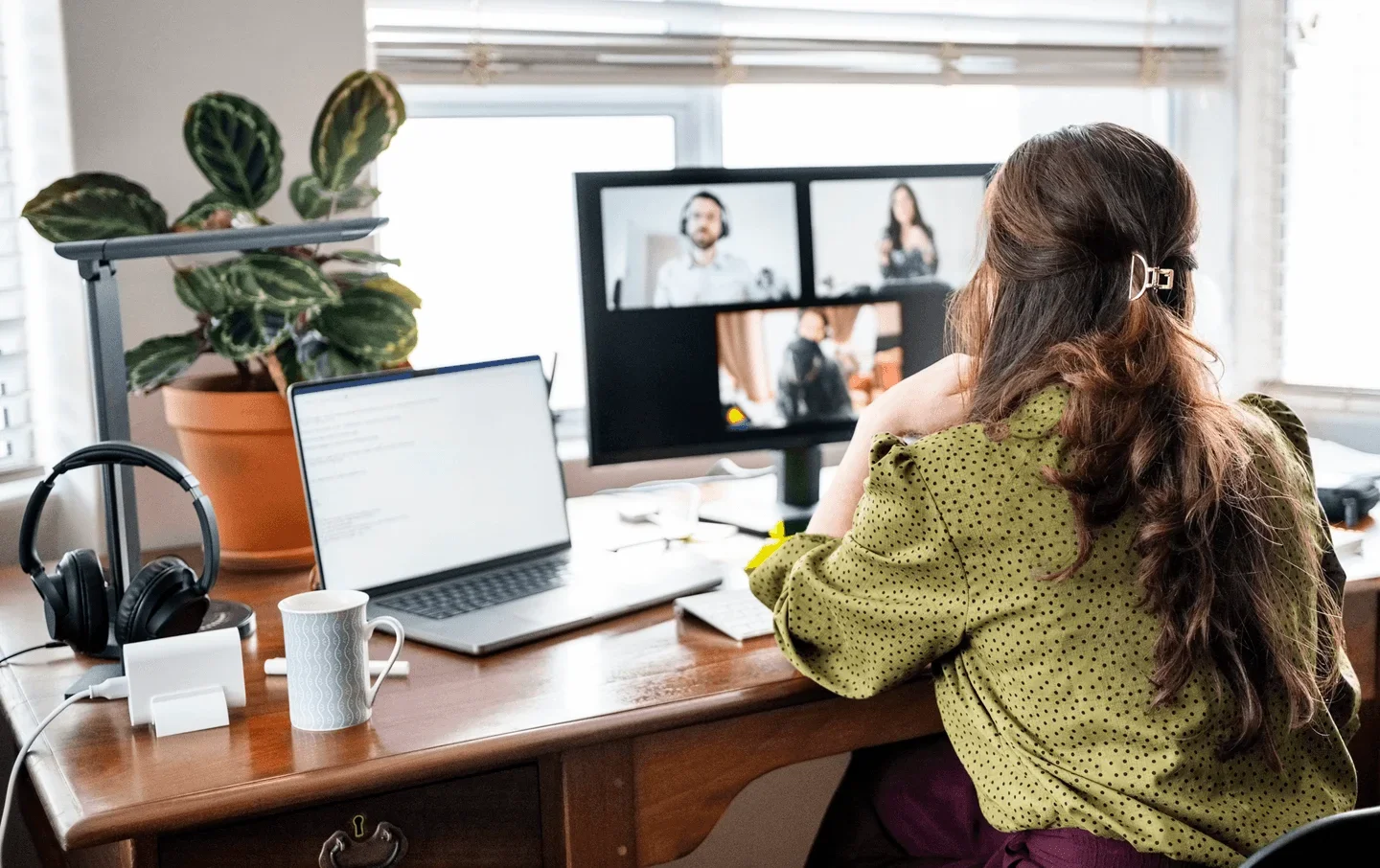 Alt text:
Person working from a home office, seated at a desk with a laptop and headset while participating in a video conference displayed on a monitor, with a coffee mug and houseplant nearby.