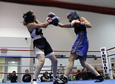 Two women in a boxing ring. They are both wearing protective gear and are mid-fight.