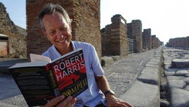 Richard E Grant smiling, and reading an open copy of the book: Pompeii