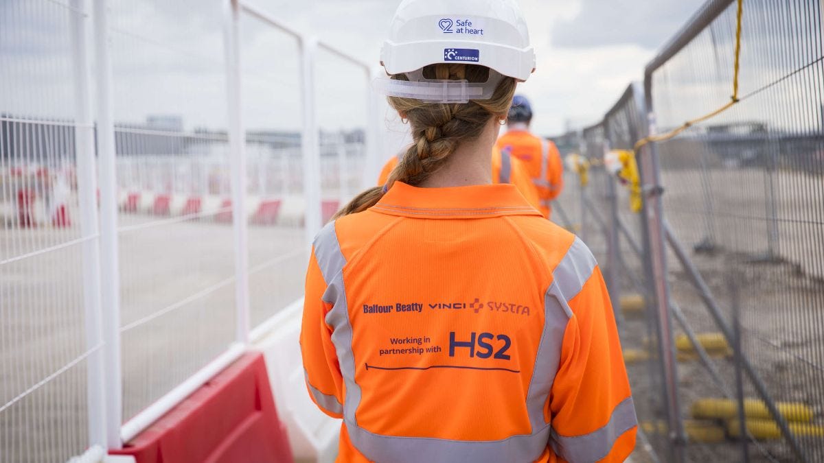 Female HS2 worker in orange overalls and hard hat walking on site.