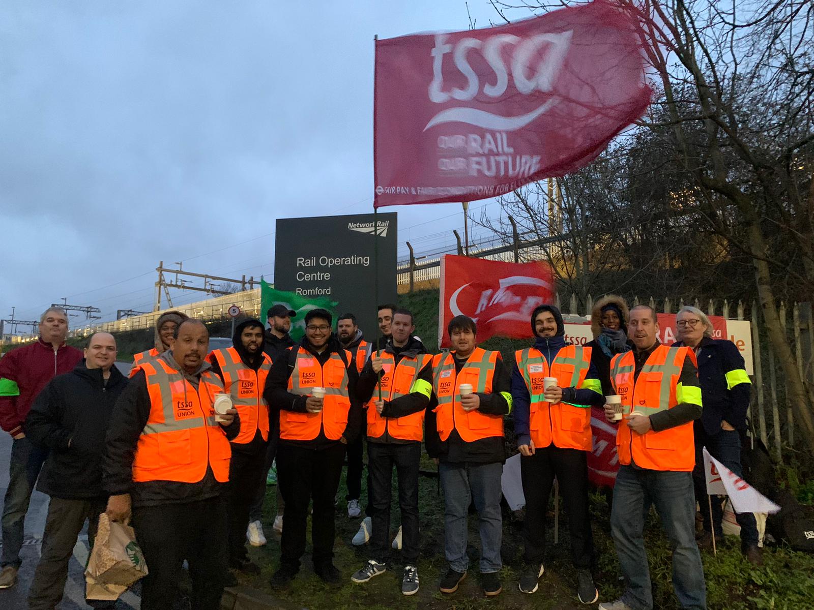A group of TSSA Elizabeth Line pickets in orang hi vis vests and yellow armbands at Romford Rail Operating Centre. A huge red TSSA flag flies above them