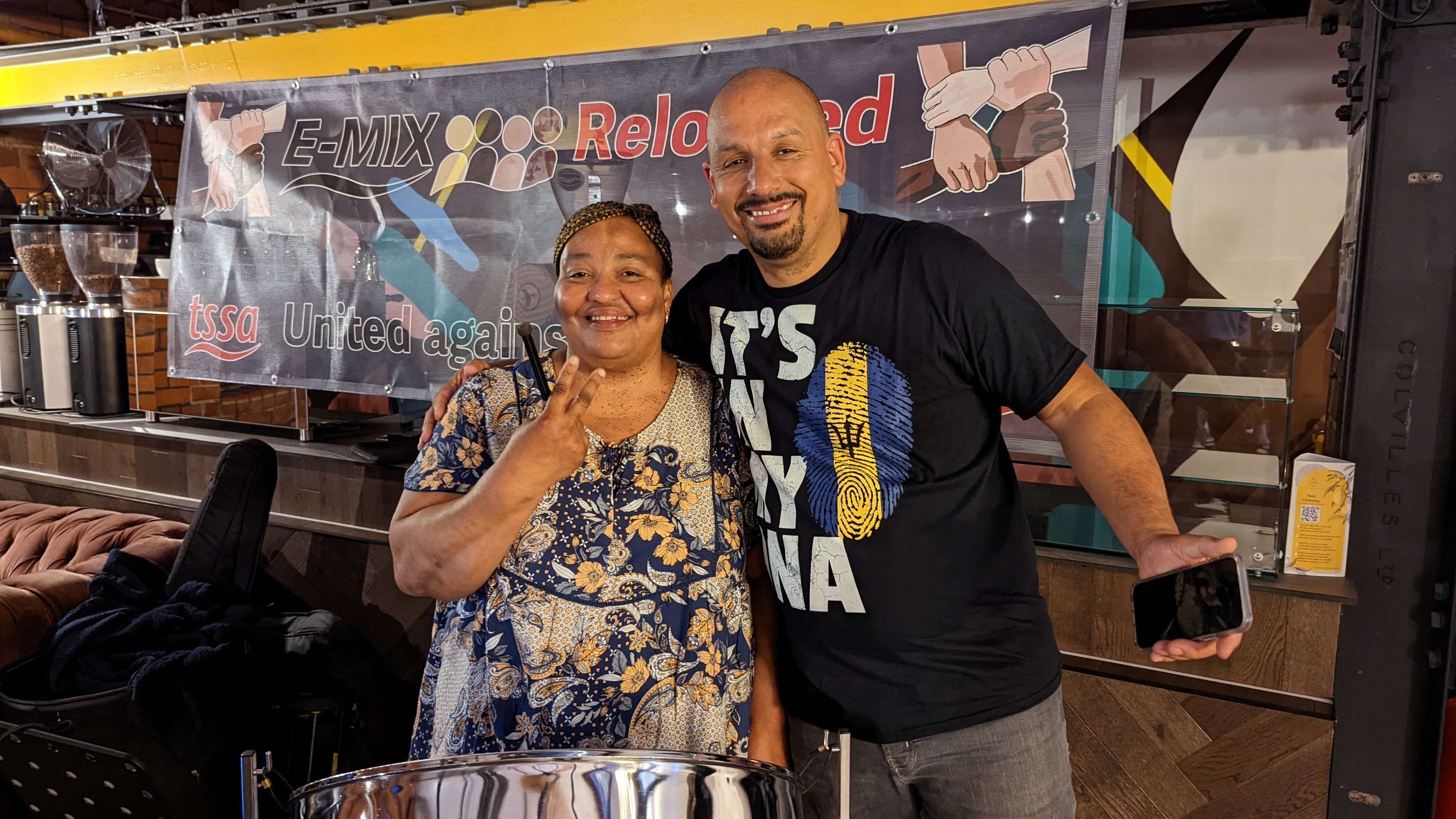 A black woman and a black man standing together. The woman is wearing a brightly covered dress and the man is wearing a black t-shirt with a blue and yellow logo. A banner behind them reads "E-mix reloaded".
