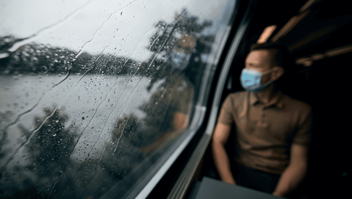 Man wearing face mask on train looking out of rain splattered window