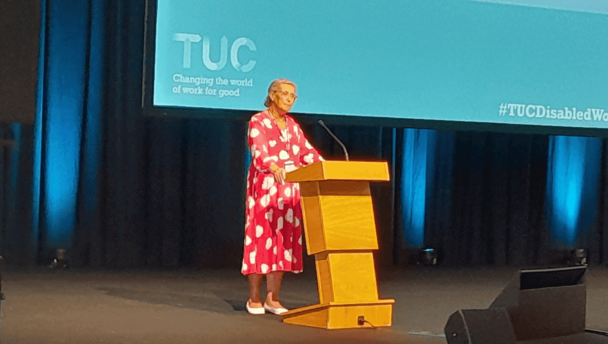 Cheryl O'Brien, a white woman with grey hair standing at a wooden podium in front of a screen saying "TUC". She is wearing a red dress with a white floral print.