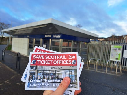 A hand holding leaflets in front of Blairhill station ticket office against a blue sky. The leaflets read "Save ScotRail Ticket Offices" and have a TSSA logo and a picture of a different ticket office on them.