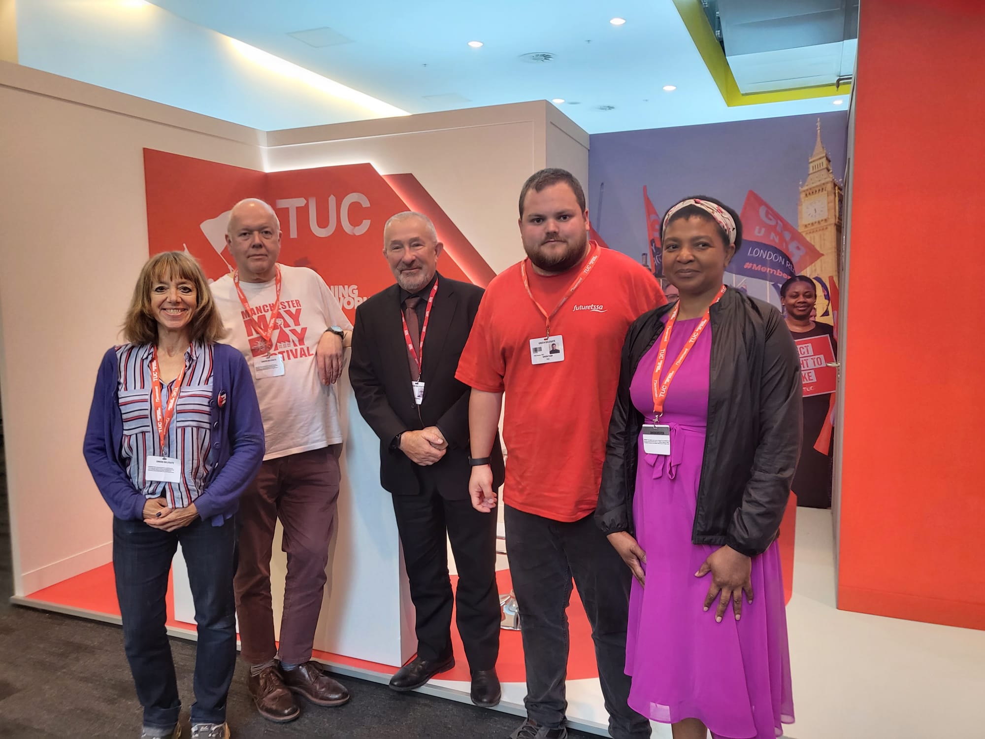 5 people stand in front of a TUC logo. A women wearing a blue cardigan, blue shirt and jeans, a middle aged man wearing a white May Day t-shirt and brown trousers, a middle aged man wearing a black suit, a younger man wearing a red tshirt and black trousers and a women wearing a purple dress and a black coat.