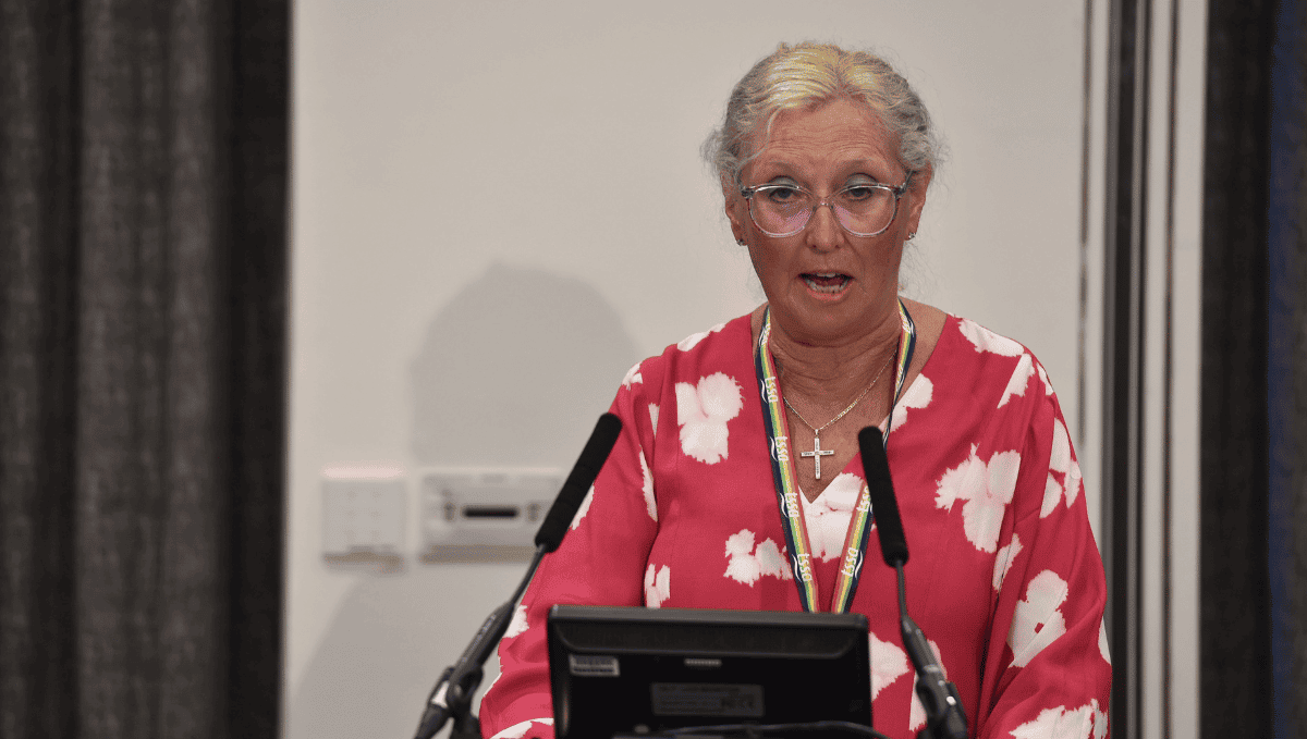 Cheryl O'Brien, an older woman in a red dress with a white print, and a gold cross. She has blonde hair, glasses and is standing at a podium.