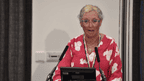 Cheryl O'Brien, an older woman in a red dress with a white print, and a gold cross. She has blonde hair, glasses and is standing at a podium.