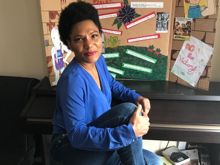 A black woman in a bright blue top and blue jeans, sitting in front of a noticeboard covered in feminist empowerment slogans.