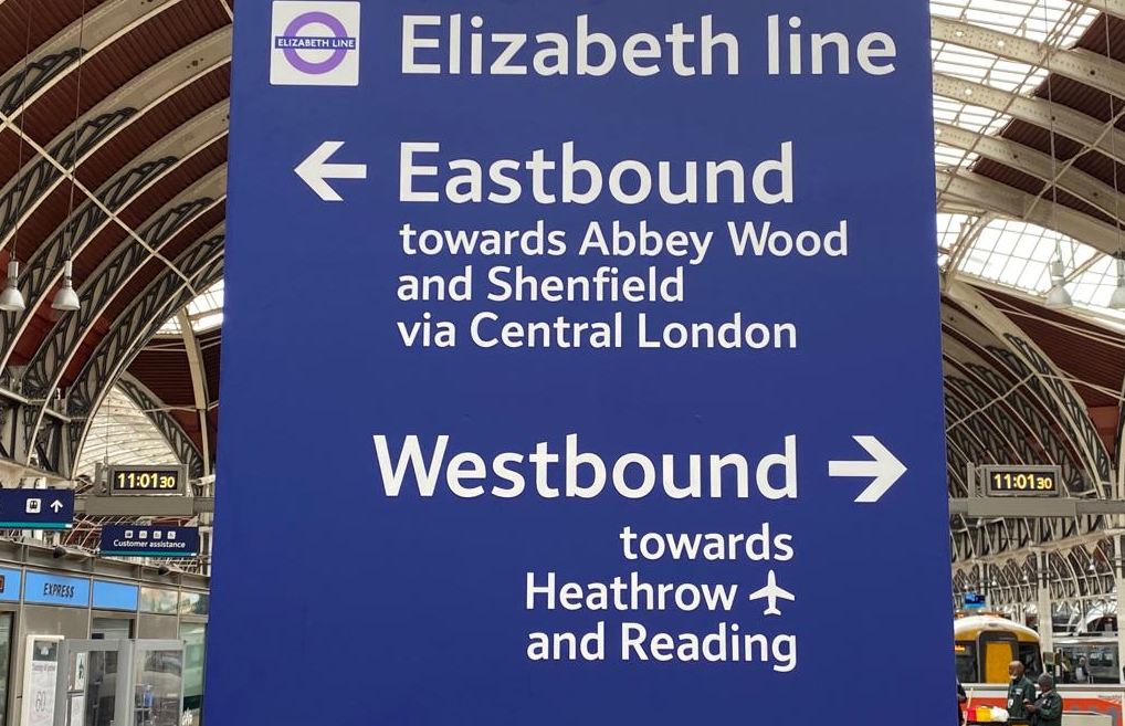 A purple Elizabeth Line sign with arrows saying Eastbound and Westbound pointing in opposite directions. Behind the sign is a mainline railway station.