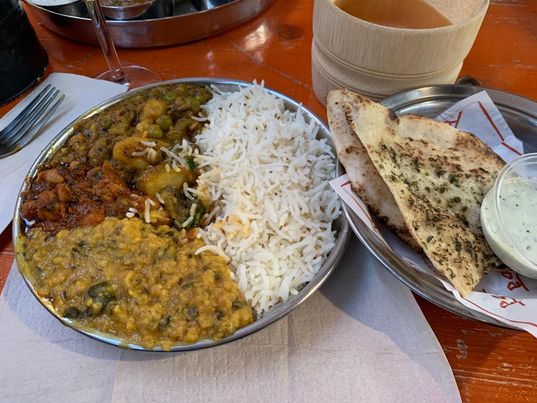 A silver circular plate with rice on one side and three different types of curry on the other. Next to it is an silver oblong plate with garlic nans and a raita dip. 