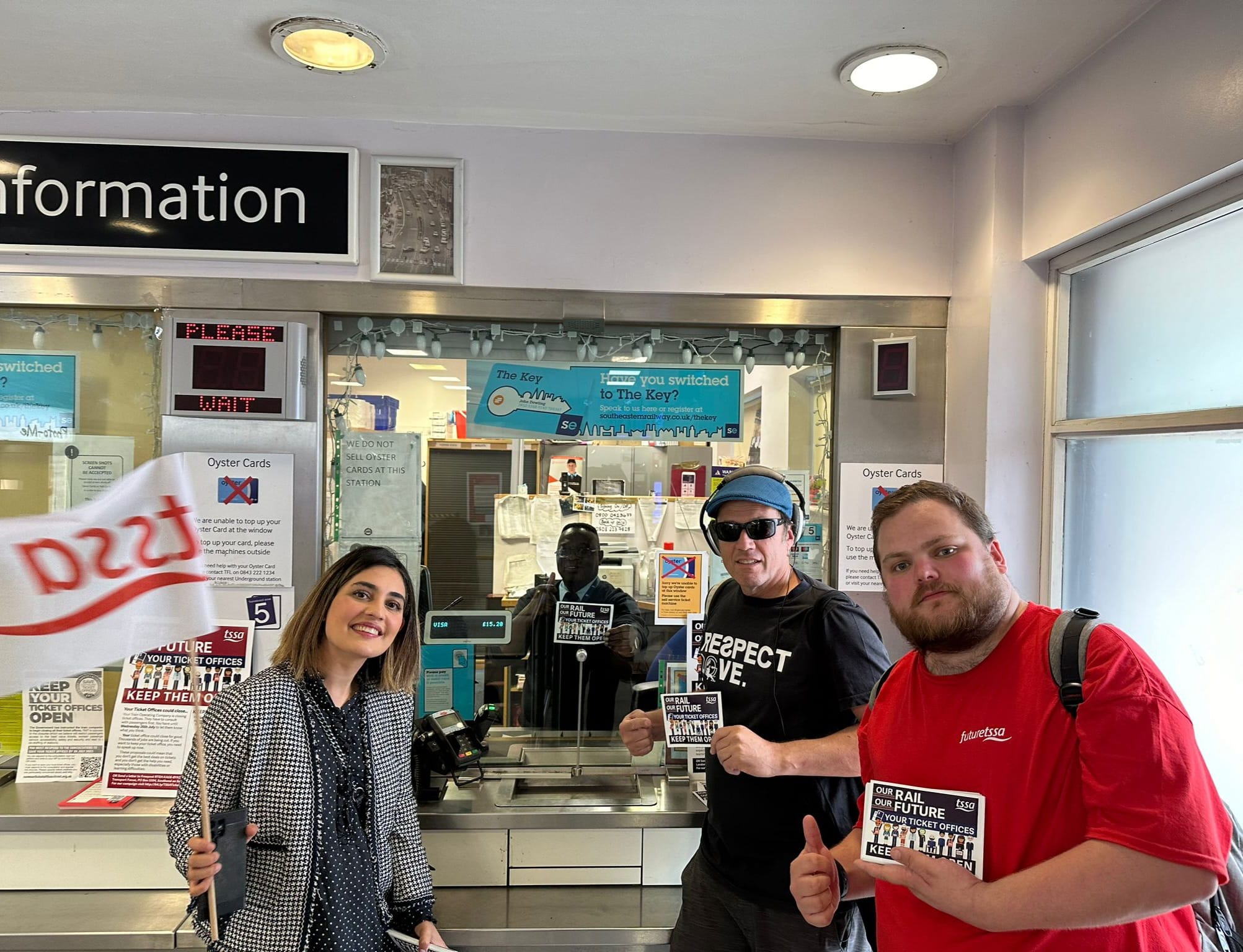 Maryam Eslamdoust, a persian woman with shoulder length hair and a grey suit holding a TSSA flag standing in front of a ticket office with a young man in a red tshirt, an older man in a black t shirt. There is a black man behind the ticket office window. They are smiling and holding TSSA ticket office campaign leaflets.