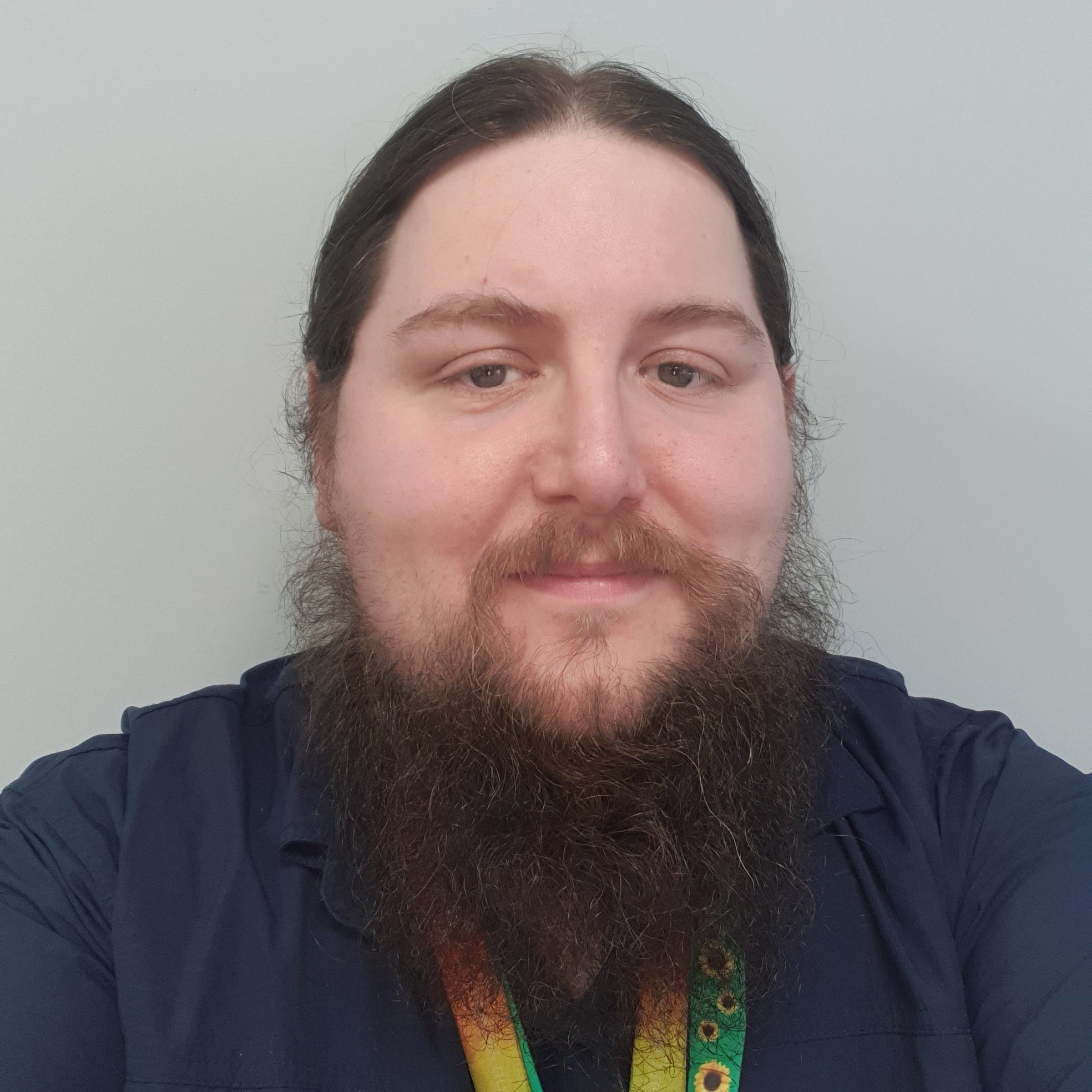 Mikey Atwell, a young white man with a large brown beard and long hair. He is wearing a dark blue shirt and a rainbow lanyard. 