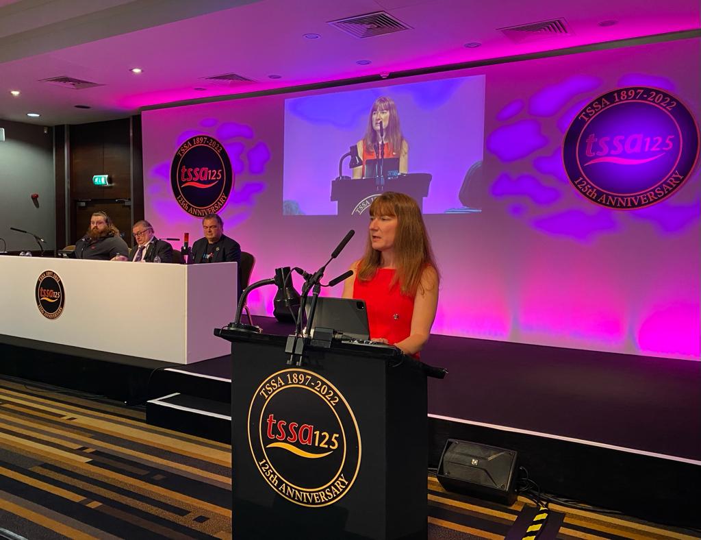 Amy Wiser, a woman with brown hair and a red dress speaks at a rostrum. Behind her are 3 men sitting at a table and a purple background. The front of the rostrum is black with a gold "TSSA 125th anniversary" logo.