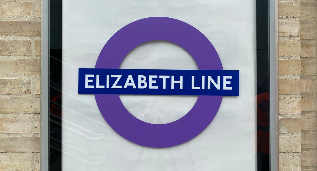 A purple Elizabeth line roundel. It is displayed on a plain white poster against a brick wall. 