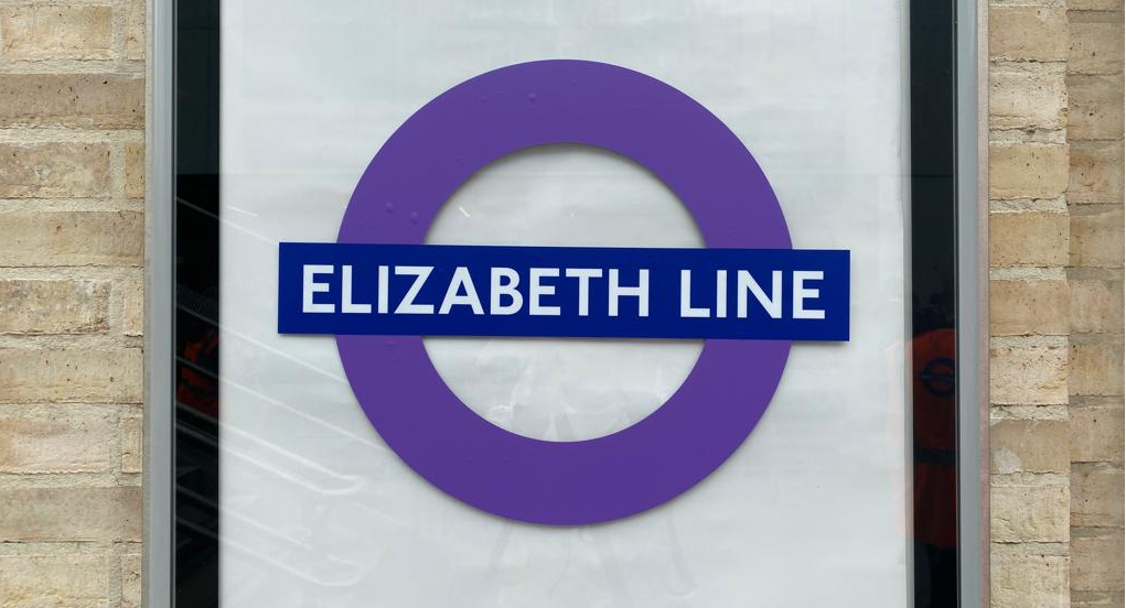 A purple Elizabeth line roundel. It is displayed on a plain white poster against a brick wall.