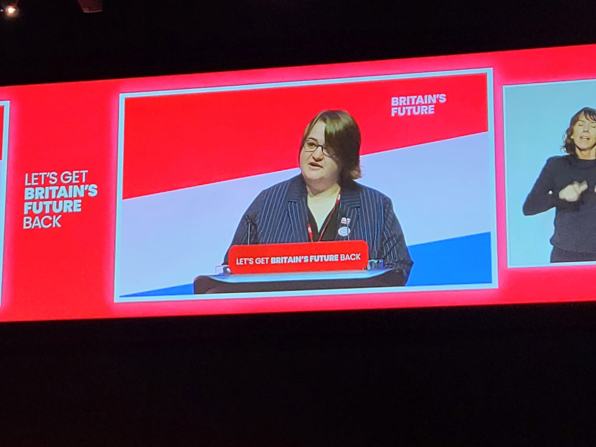 TSSA President Melissa Heywood, a white woman with brown hair, wearing a dark pinstriped shirt, stands at a podium speaking to Labour conference. Behind her are red, white and blue stripes on the wall. On one side is a sign language translator. 