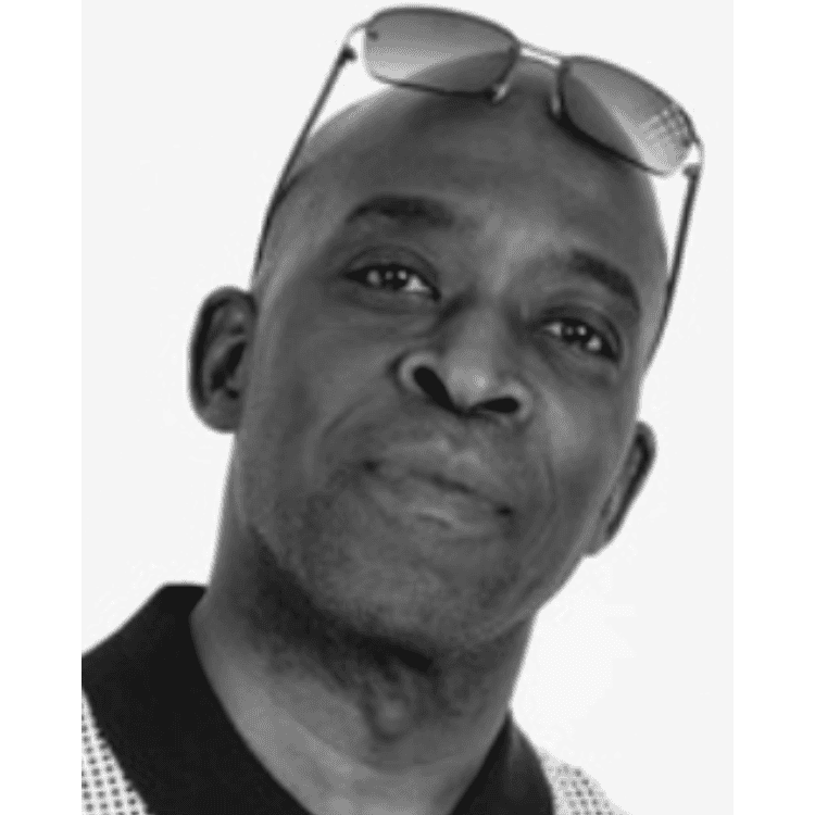 A black-and-white photo of Abdul Ajayi, a bald black man with sunglasses resting on his forehead. He is wearing a spotty shirt with a black collar and looking calmly at the camera.