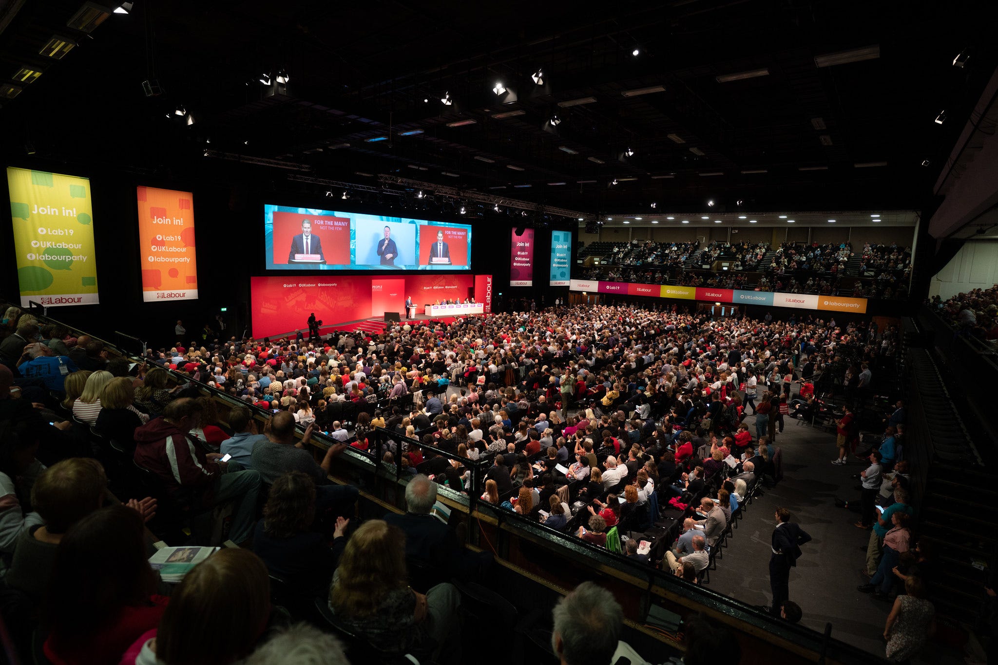 Picture of Labour Party conference hall, Brighton 2019, showing rows of seated delegates, stage and big screens