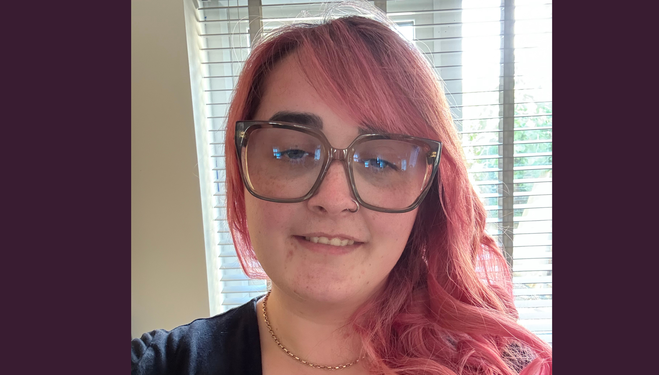 Honor Travers, a white woman in a black t-shirt looking at the camera. She has large glasses and long pink hair. Behind her is a window with sunlight shining through closed blinds.