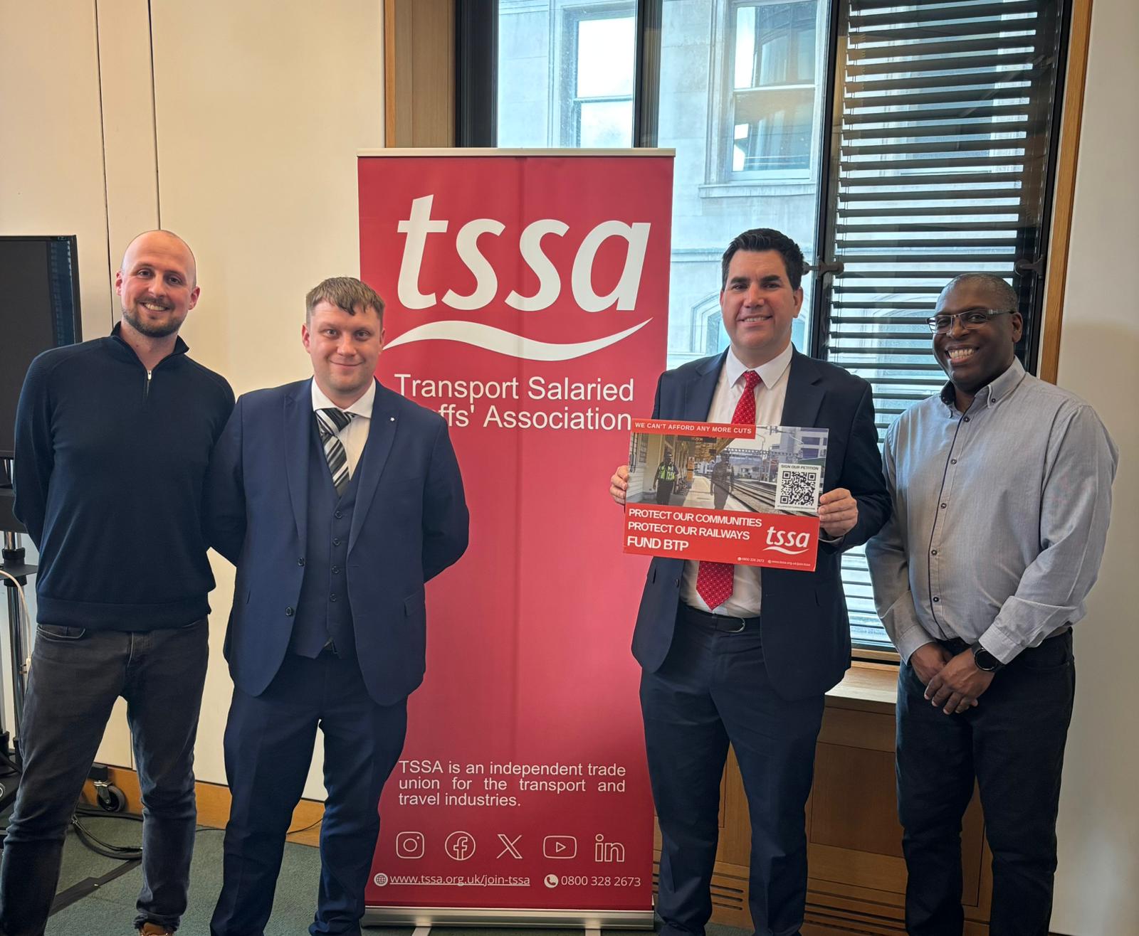Four men standing in a line side by side. Red TSSA banner in the background. One of the men, Richard Burgon MP, is holding a sign calling for an increase in funding for the British Transport Police.
