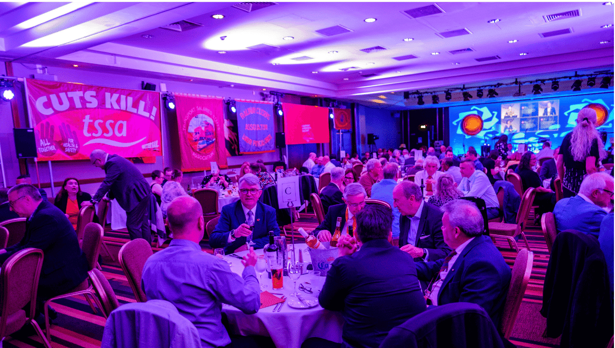 A busy room full of people sitting at round tables, chatting together with lots of political TSSA flags on the wall. 