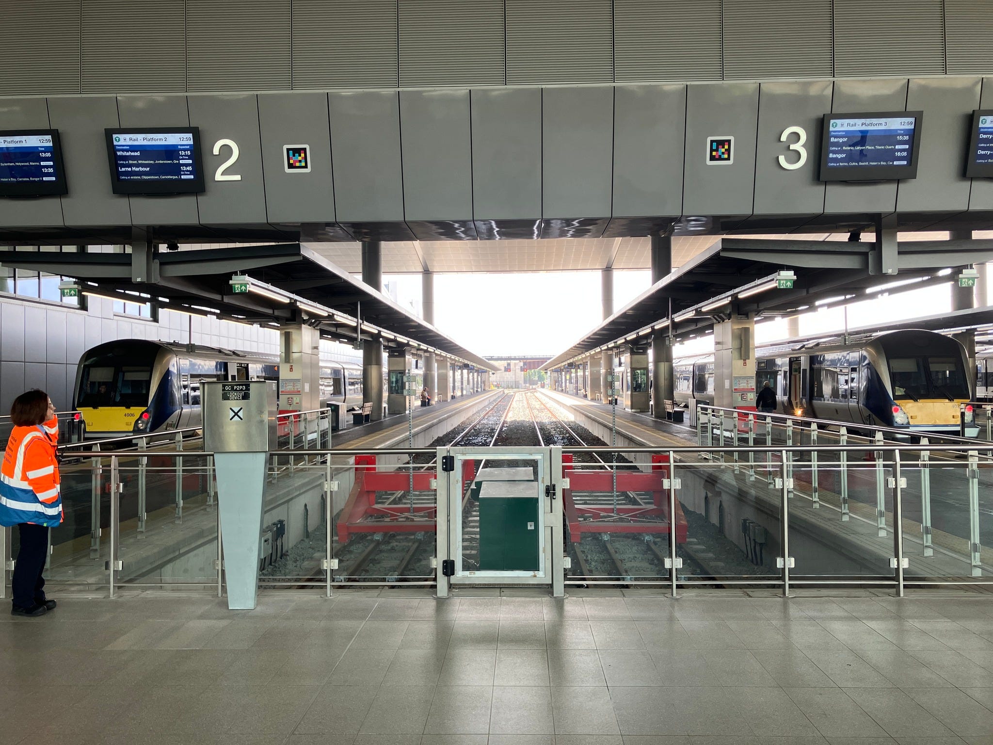 Two trains standing at platforms at Belfast train station.Two empty platforms lie between them. There is a departures board above the platforms, labelling the empty ones as Platform 2 and 3. Someone in a hi vis jacket is standing on the left of the platform.
This station is part of the Translink network.
photo credit: Alexandra Taylor, used with permission.