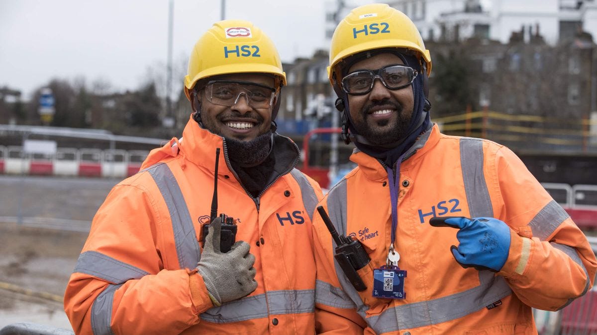 Two BAME HS2 workers in orange overalls, yellow hard hats and safety specs on site with radios.