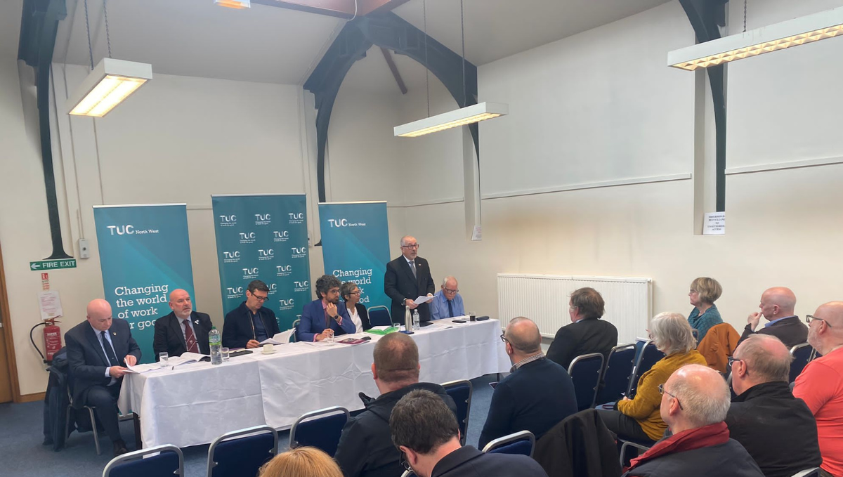 Peter Pendle, a middle aged bald white man with glasses stands, speaking behind a table with other people sitting at it. The audience faces him and we can see the backs of their heads. Also sitting at the table are Andy Burnham, Mayor of Manchester, Mick Whelan, leader of ASLEF an Mick Lynch leader of RMT.  