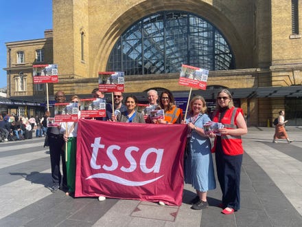 Maryam Eslamdoust, Maria Fawcett and TSSA staff and activists gathered around a huge TSSA banner in front of Kings Cross station. They are holding placards campaigning for British Transport Police to get better funding. The sky is bright blue, and it is bright and sunny.  