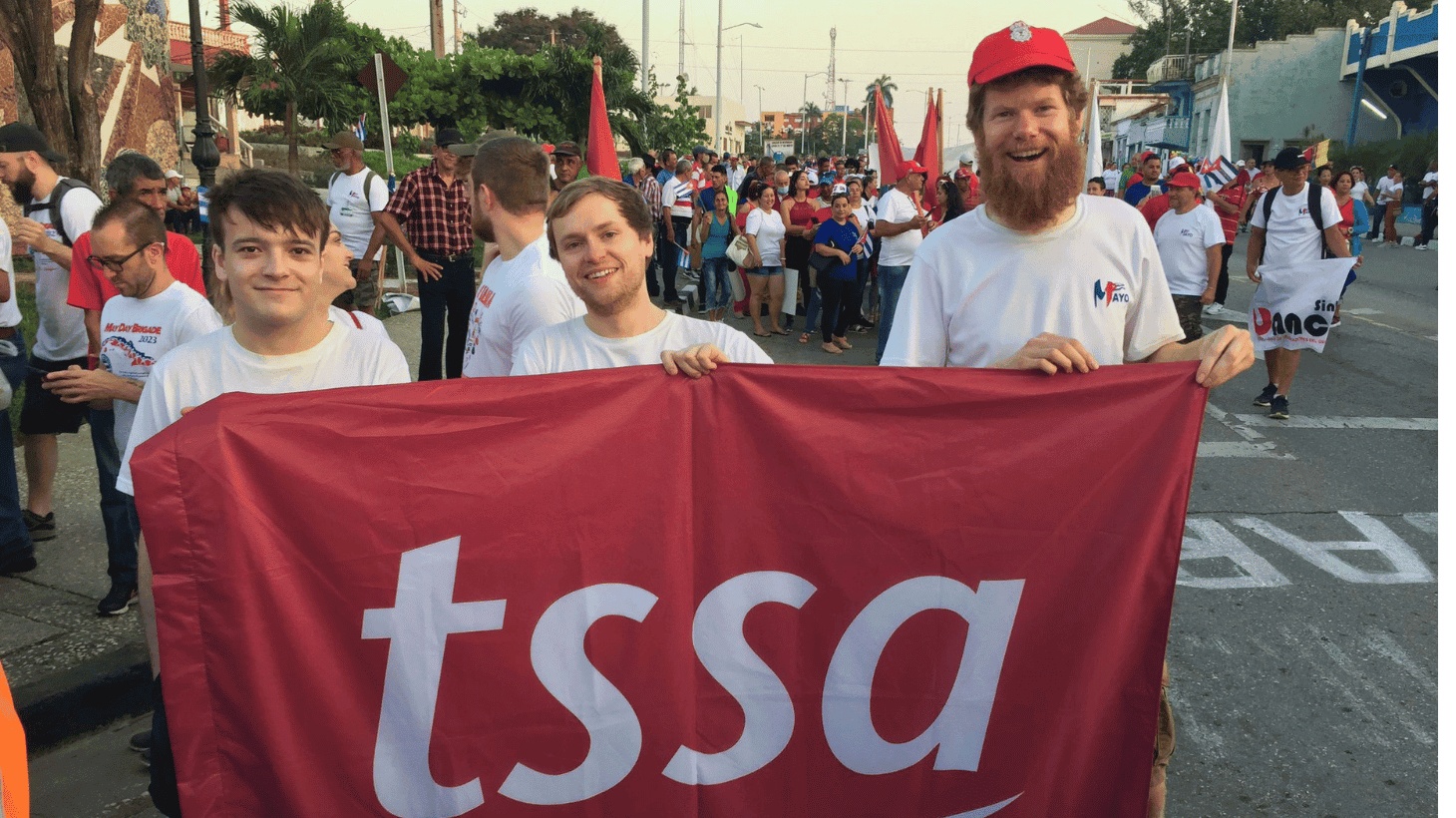 Three young men, holding a huge red TSSA  flag, and smiling happily on a street in Cuba. Will Boisseau with his big red beard, wearing a red baseball cap is on the right of the flag.