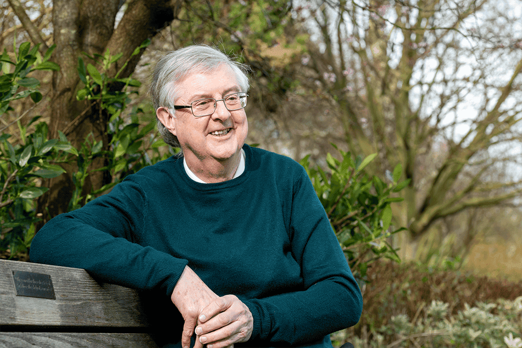 Mark Drakeford, an elderly Welsh man, with white hair and glasses,wearing a green sweater. He is sitting on a wooden bench, looking off to the side and smiling. There are trees in the background.